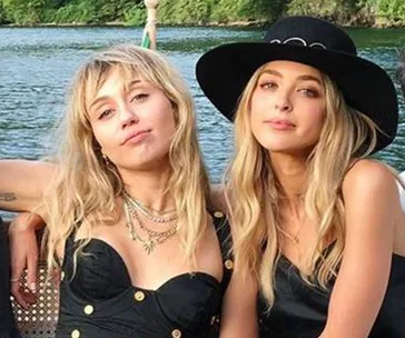 Two women sitting together outdoors by a lake; one wears a black dress, the other a hat.