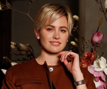A person with short blonde hair in a brown jacket poses in front of floral branches.