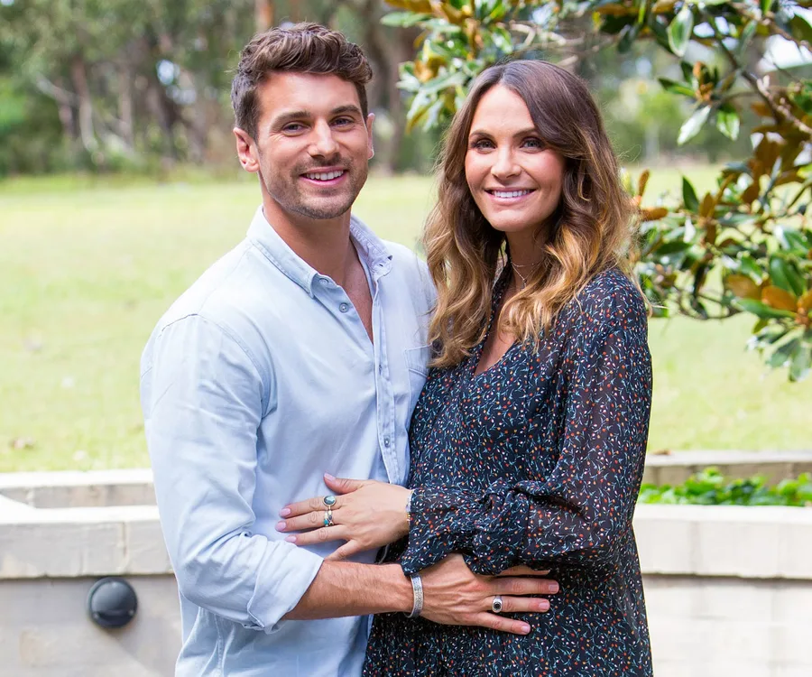 A couple smiling and embracing in an outdoor setting with greenery in the background.