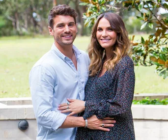 A couple smiling and embracing in an outdoor setting with greenery in the background.