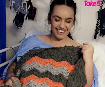 A woman in a blue hospital gown with medical equipment, smiling and holding a crochet blanket.