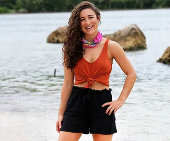 A person in a rust top and black shorts stands smiling on a beach with rocks and water in the background.