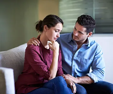 A man comforts a sad woman on a couch, gently holding her shoulder and hand, both wearing casual clothes in a modern room.