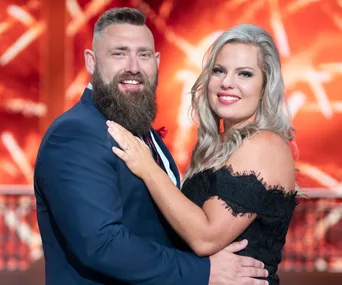 A smiling couple embraces, with the woman in a black dress and the man in a suit, set against a warm, blurred red background.