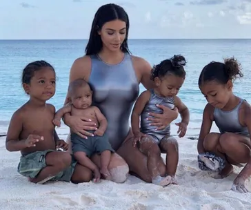 Woman in grey swimsuit on the beach with four children, two boys and two girls, playing in the sand.
