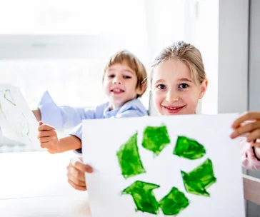 Children holding drawings of recycled symbols, smiling indoors, promoting environmental awareness.