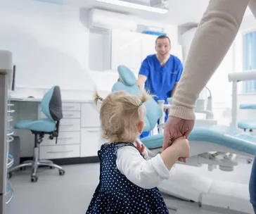 A toddler holding an adult's hand in a dental clinic while a dentist in blue scrubs smiles in the background.