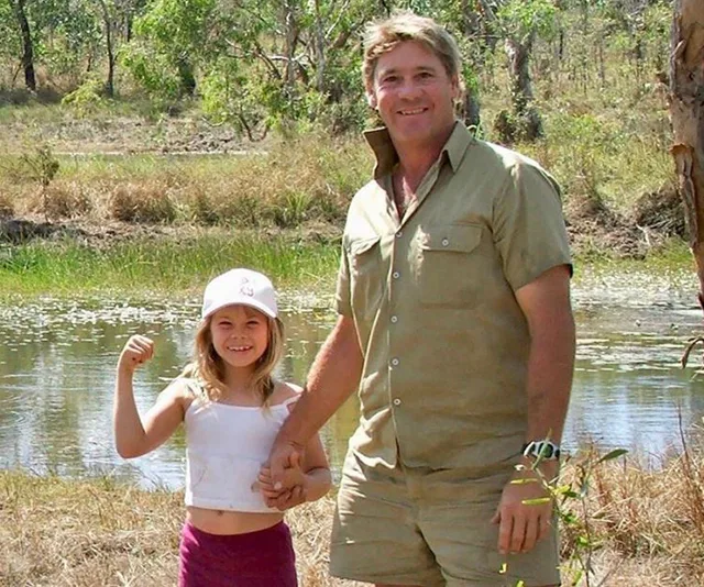Father and daughter smiling by a pond in the wilderness, with the girl flexing her arm.