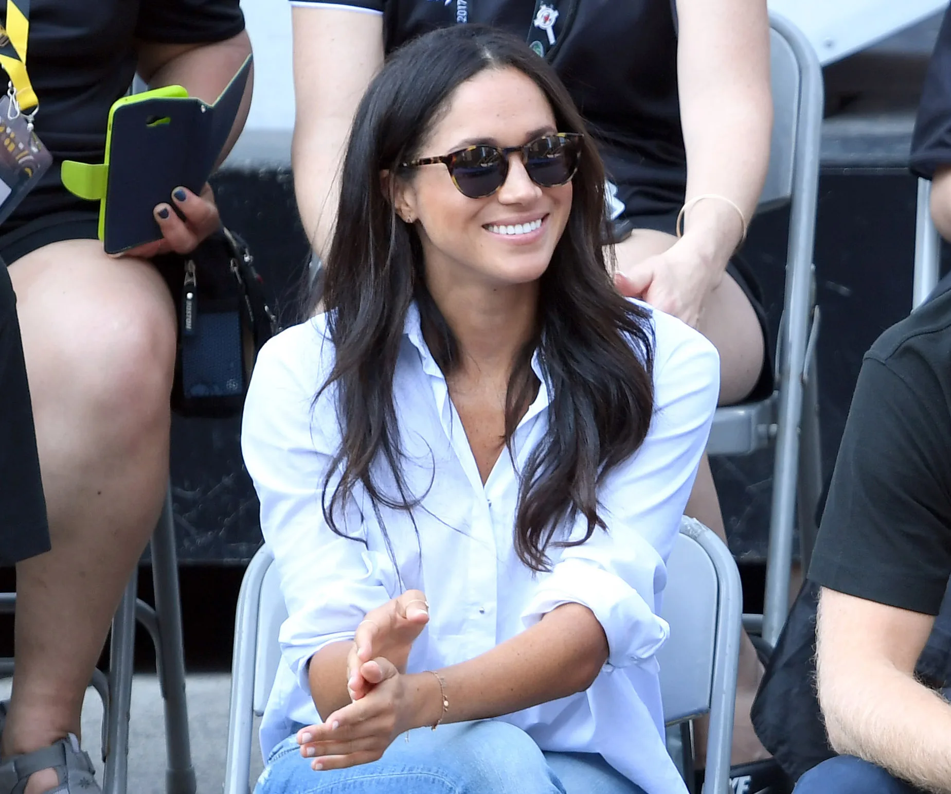 A woman in sunglasses and a white shirt smiles while seated outdoors at an event.