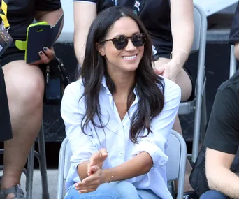 A woman in sunglasses and a white shirt smiles while seated outdoors at an event.