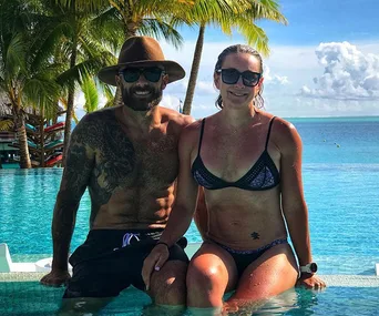 Man and woman in swimsuits sitting by a pool, with palm trees and ocean in the background.