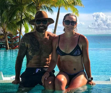 Man and woman in swimsuits sitting by a pool, with palm trees and ocean in the background.