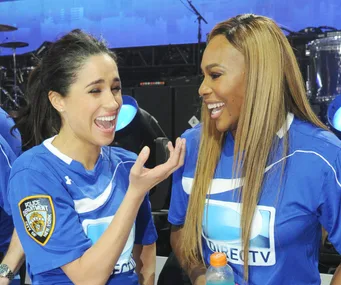Two women in blue sports jerseys laugh together, standing indoors in front of a stage with musical instruments.
