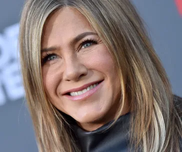 Smiling woman with long, highlighted hair at an event, wearing a black outfit on a gray background.