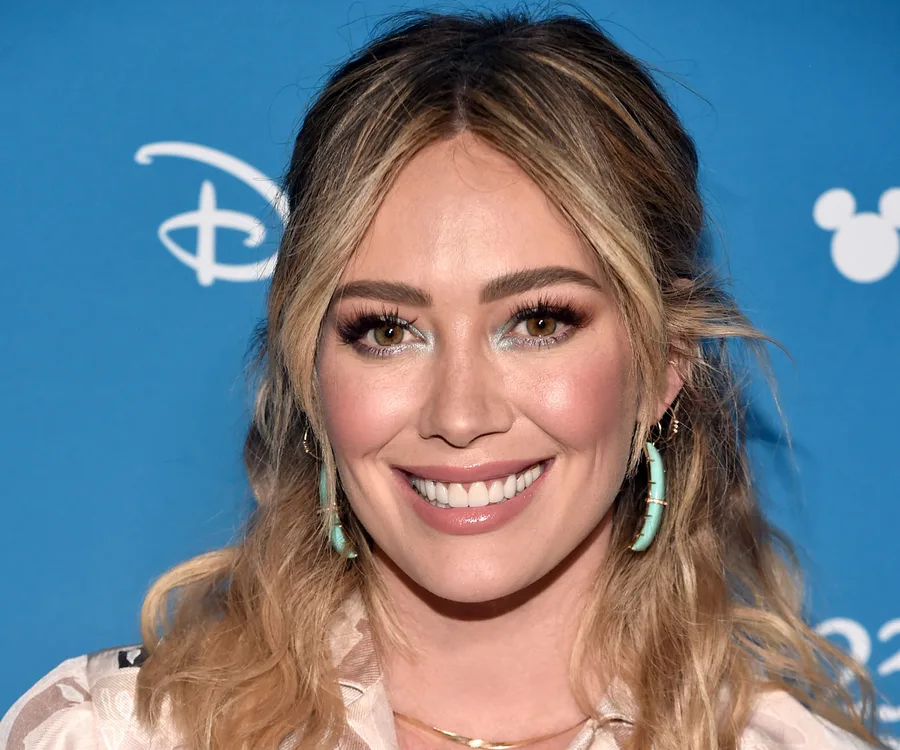 A woman smiling at a Disney event, wearing turquoise earrings and a white top, with a blue background.