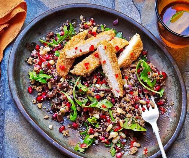 Breaded fake chicken slices on quinoa salad with greens and pomegranate seeds, beside an orange drink.