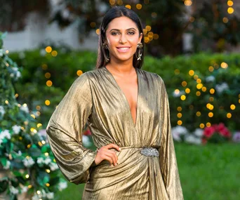 Woman in a gold dress posing outdoors with a backdrop of greenery and twinkling lights.