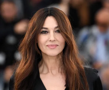 Monica Bellucci at a public event, smiling softly, with long, wavy brown hair against a blurred background.