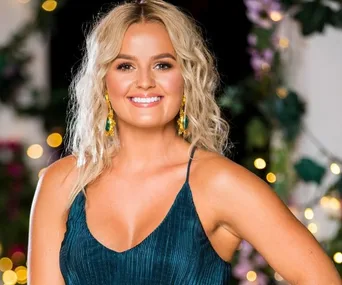 A woman with wavy blonde hair in a blue dress smiles in a garden setting with lights and greenery.