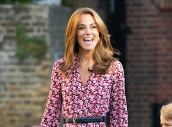 Kate Middleton in a floral Michael Kors dress, smiling outdoors with a brick wall background.