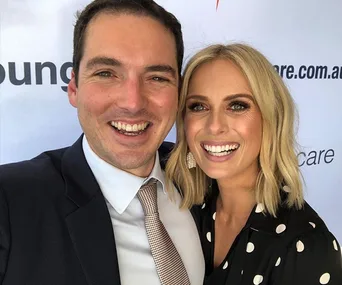 Two people smiling, dressed formally, standing close to a backdrop with partial text.