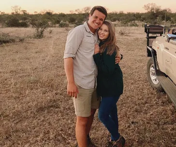 A couple poses happily in a grassy open field near a safari vehicle at sunset.