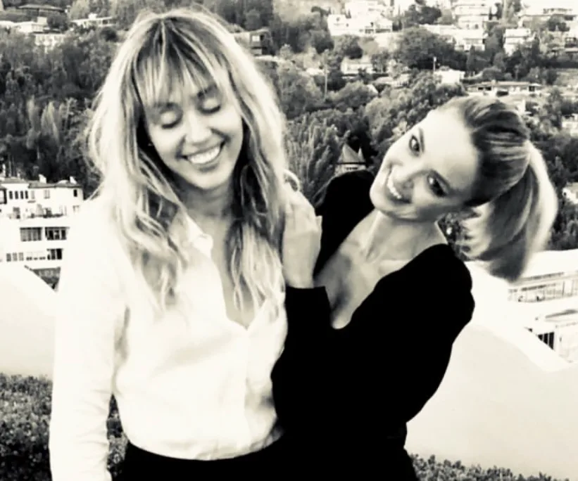 Two women smiling outdoors with a cityscape in the background, one with fringed hair and the other in a ponytail.