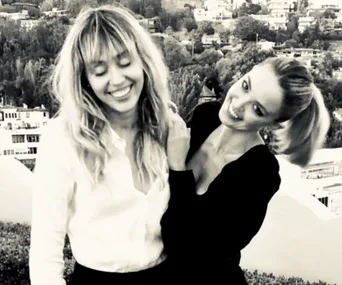Two women smiling outdoors with a cityscape in the background, one with fringed hair and the other in a ponytail.
