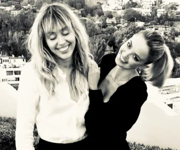 Two women smiling outdoors with a cityscape in the background, one with fringed hair and the other in a ponytail.