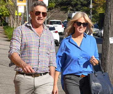 Couple walking outdoors on a sunny day, both wearing sunglasses, he in a checkered shirt, she in a blue blouse, carrying a bag.