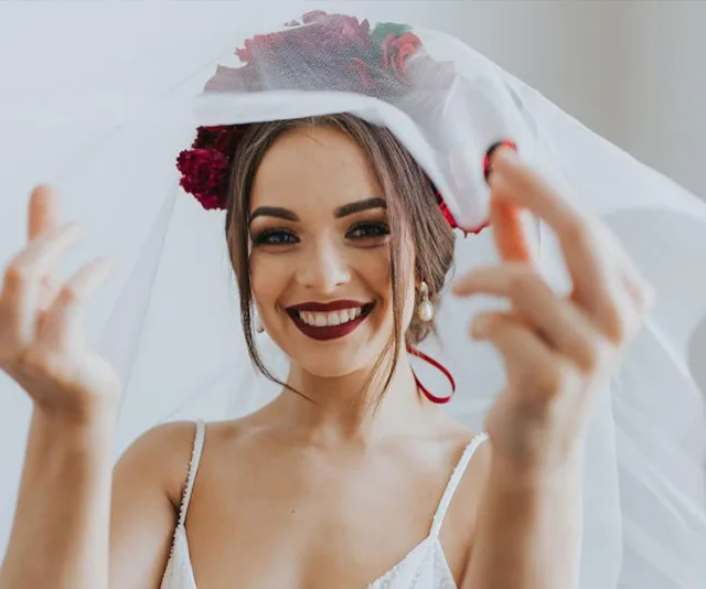 Smiling bride in white dress with veil and flower crown, adjusting her veil.