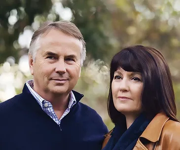 A couple standing outdoors, dressed in fall clothing, with blurred greenery in the background.