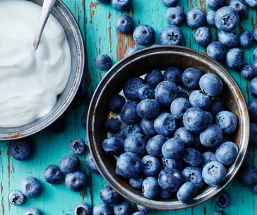 Bowl of fresh blueberries next to a bowl of white yogurt with a spoon on a rustic turquoise wooden table.