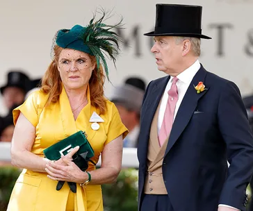 Man in formal suit and top hat stands beside woman in bright yellow outfit and green hat at a formal event.