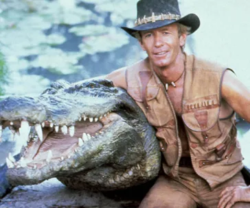 Man in a leather vest with a crocodile prop, wearing a black hat, set outdoors.