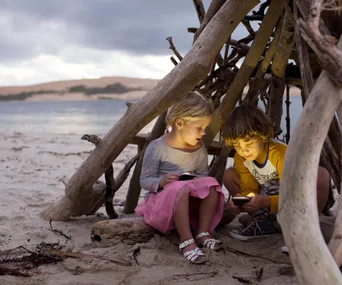 Children sitting under a makeshift driftwood shelter on a beach, illuminated by their handheld devices.