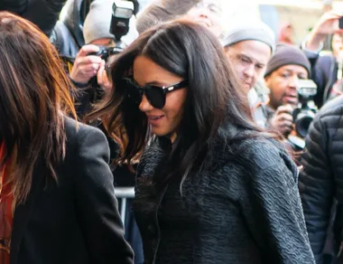 A woman with sunglasses and dark hair walks through a crowd, surrounded by photographers.
