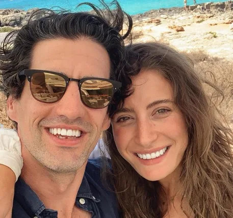 A smiling couple takes a selfie on a sunny rocky beach with turquoise water in the background.