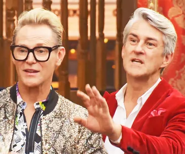 Two men are standing indoors. One has short blond hair and glasses, wearing a glittery jacket; the other with gray hair in a red blazer.