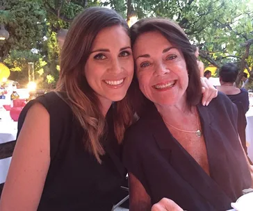 Two women smiling at an outdoor dinner setting, surrounded by trees and soft lighting.