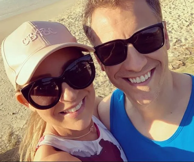 Couple wearing sunglasses and smiling at the beach with the sand and sea in the background.