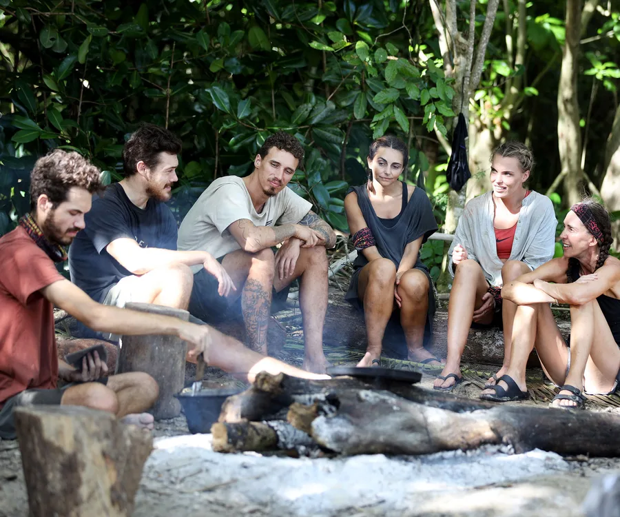 Group of six "Survivor Australia" 2019 contestants sitting by a campfire in a tropical forest setting.