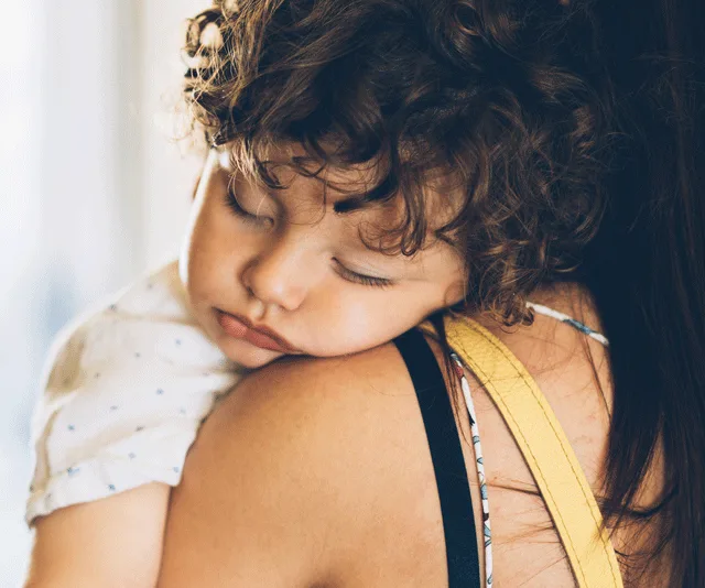 A sleeping toddler with curly hair rests on a woman's shoulder, wearing a light-colored shirt.