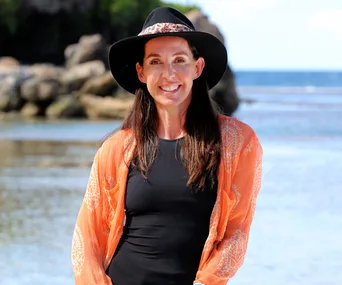 Woman in a black hat and orange shawl, smiling on a beach with rocks and ocean in the background.