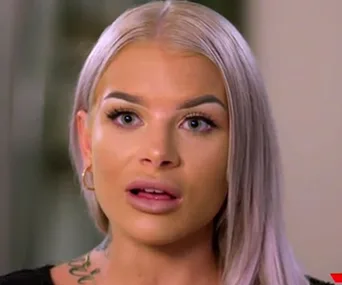 A woman with light blonde hair and tattoos, looking surprised or concerned.