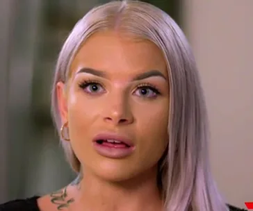 A woman with light blonde hair and tattoos, looking surprised or concerned.