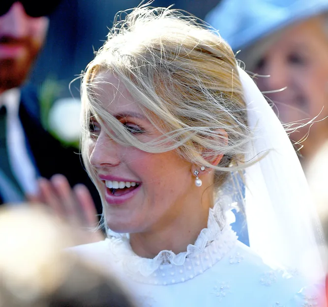 Bride with veil and pearl earrings, laughing joyfully outdoors with people in the background.