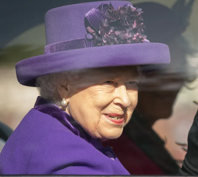 Queen in a purple outfit and hat with flowers, smiling inside a vehicle at the Braemar Games 2019.