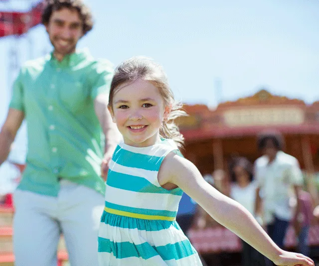 A smiling young girl in a striped dress holds an adult's hand, with a colorful amusement park in the background.