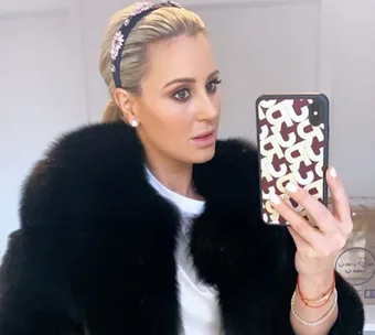 Woman taking a mirror selfie wearing a dark fur coat, floral headband, and holding a patterned phone case.